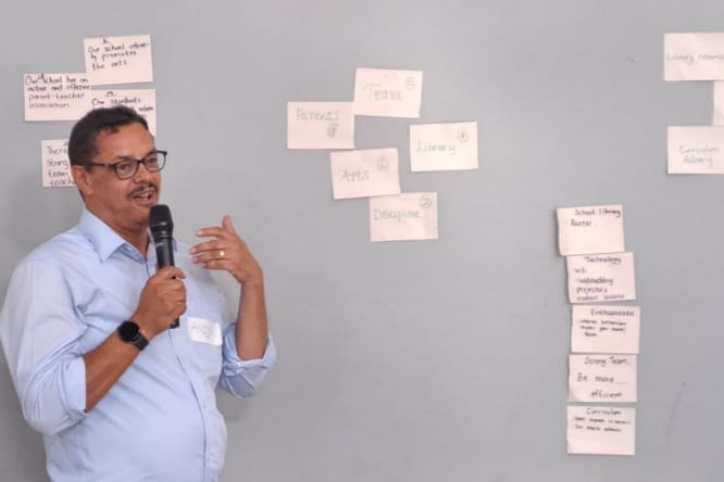 A man speaking beside a wall of handwritten cards grouping the school’s priorities: parents, team, arts, library and discipline