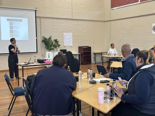 A Dinaledi coach presenting the GROW coaching model to educators seated at tables in a school hall