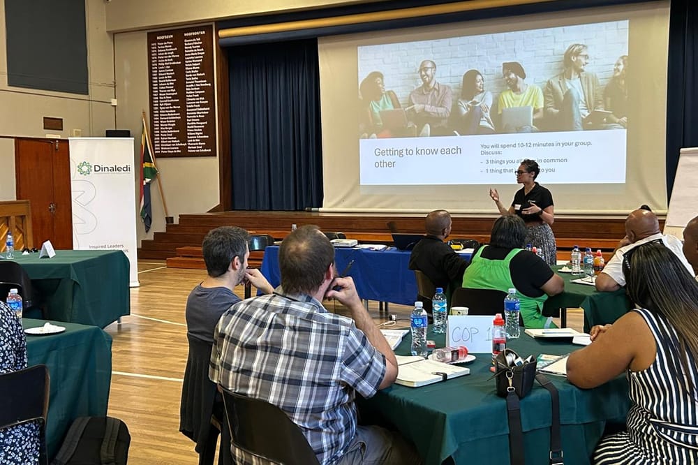 Educators at group tables in a school hall while a Dinaledi coach facilitates from the front, the Dinaledi banner beside the stage